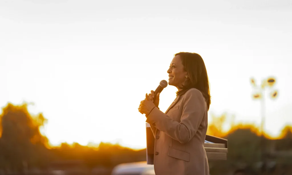 Kamala Harris against a bright outdoor background holding a microphone.