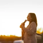 Kamala Harris against a bright outdoor background holding a microphone.